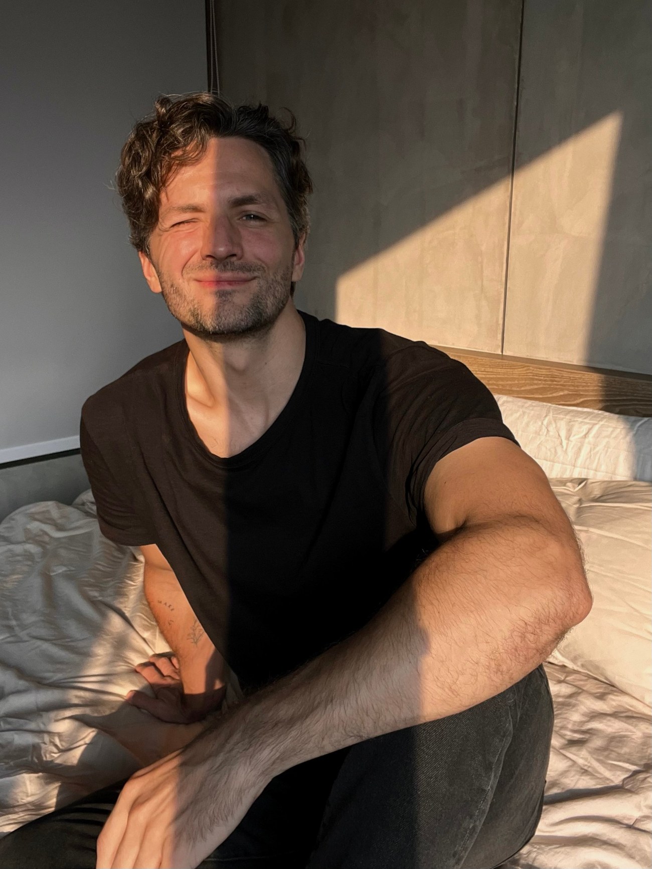 Nick Yuryev in a black T-shirt sitting on a bed in low evening sun, smiling