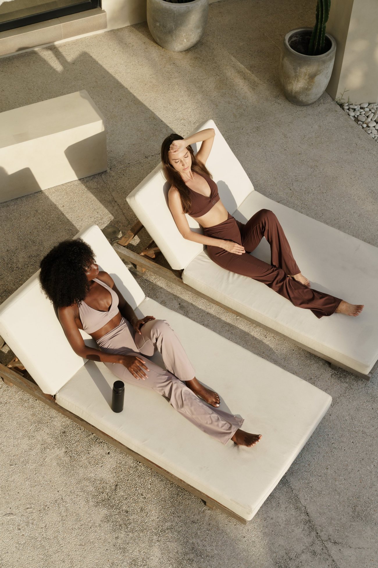 Two women in AYO sets resting on white loungers, seen from above in low sun