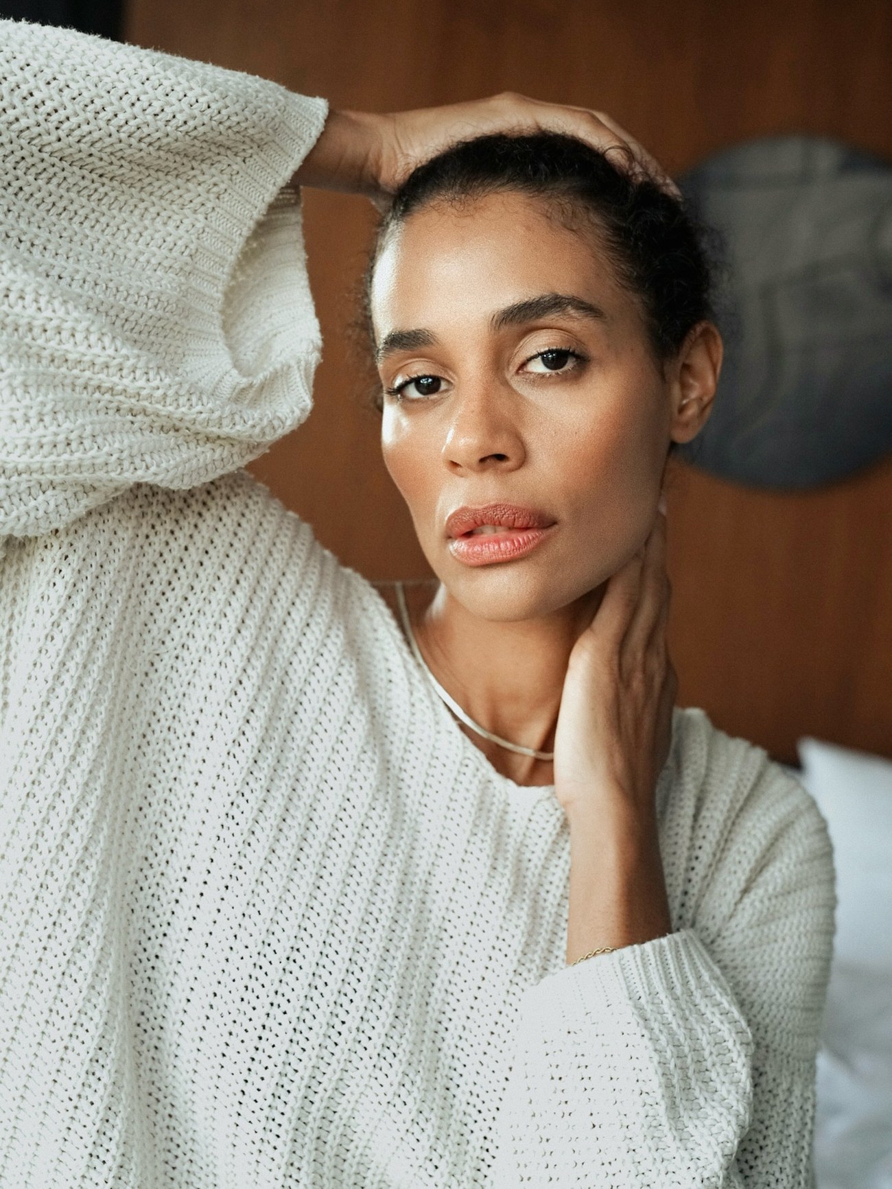 Carole in a chunky white knit sweater, hand on her neck, against a wooden wall