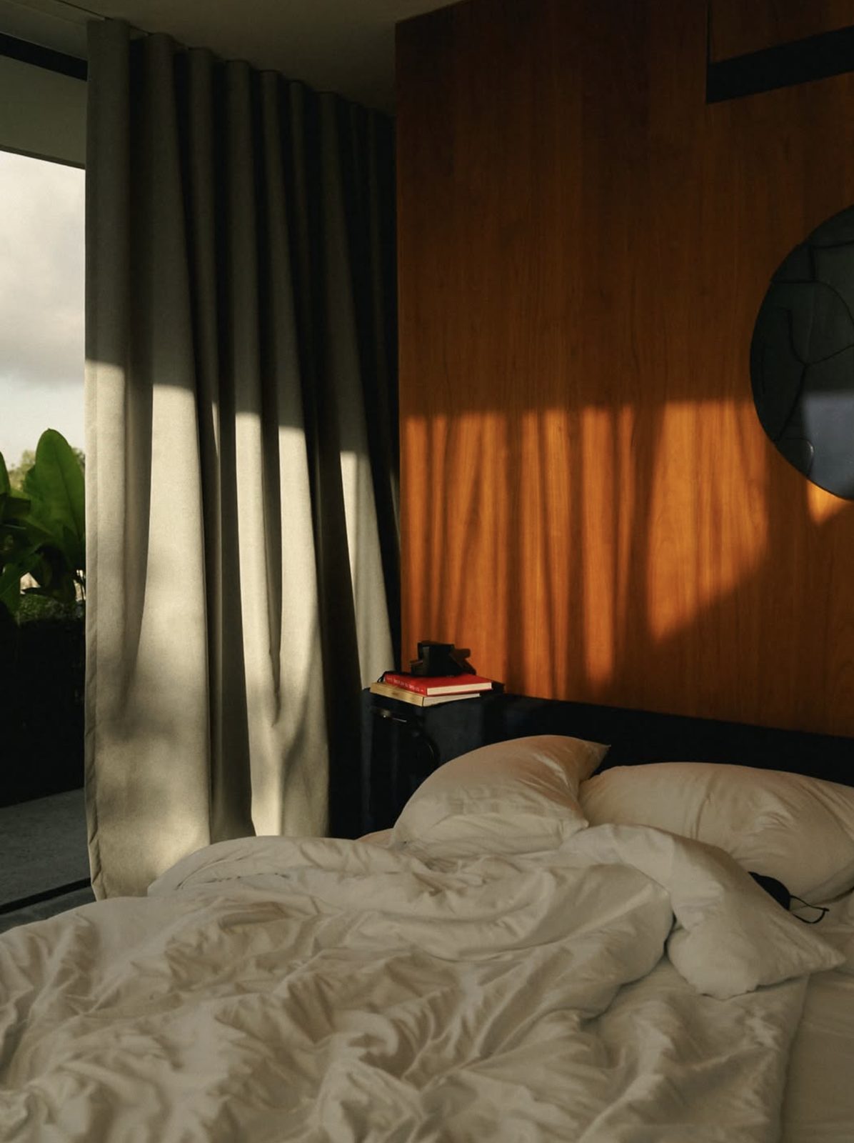 Unmade bed with low sun striping the curtain and a wooden wall