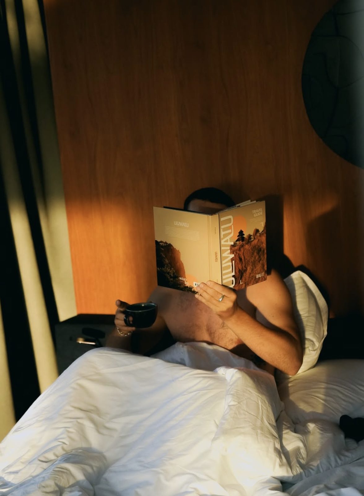 Guest in bed hiding behind an Uluwatu travel guide, coffee in hand